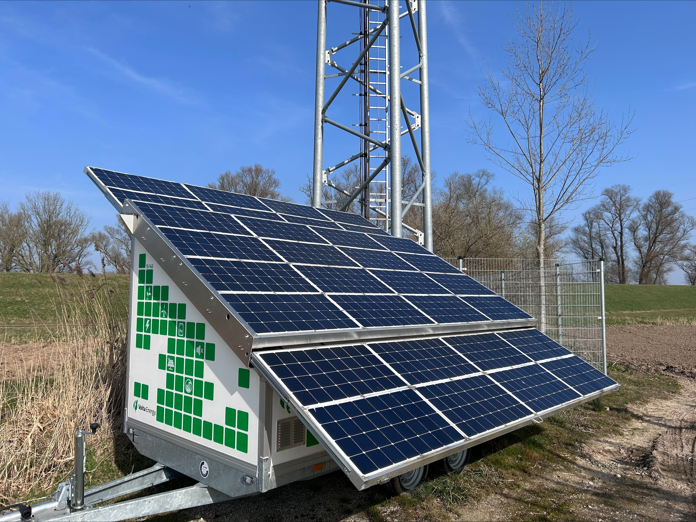 A mobile site on a lattice tower in a field. Parked in front of the site is a 2-axis trailer outfitted with 16 photovoltaic panels that power the equipment.
